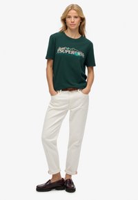 Kobieta z blond włosami w leśnozielonym t-shircie z grafiką, kremowych spodniach z podwiniętymi nogawkami, brązowym pasku i ciemnych skórzanych mokasynach, stojąca z rękami w kieszeniach.