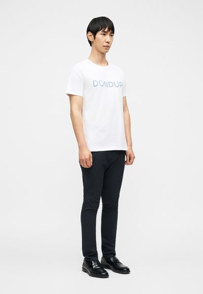 Άνδρας που φοράει λευκό μπλουζάκι με το κείμενο "DONDUP", μαύρο στενό παντελόνι και μαύρα επίσημα παπούτσια, στέκεται μπροστά σε απλό ανοιχτό φόντο.