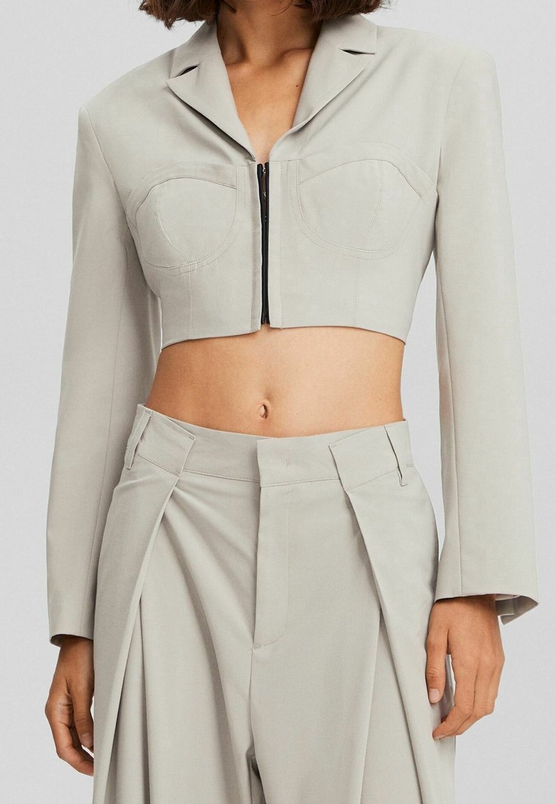 Blazer beige court à manches longues avec fermeture éclair devant, associé à un pantalon beige plissé taille haute.
