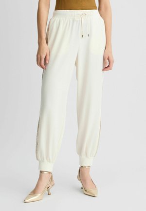 Vrouw draagt witte joggerbroek met elastische boorden en pailletten zijstrepen, gecombineerd met gouden slingback pumps met een puntige neus.