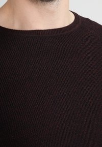 Zbliżenie na osobę w ciemno brązowym, teksturowanym swetrze dzianinowym, ukazujące szyję i górną część klatki piersiowej.