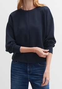 Mörkblå sweatshirt med avslappnad passform, rund hals och muddade ärmslut. Tillverkad av ett mjukt, strukturerat tyg. Kombinerad med blå jeans.