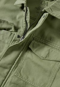 Olivově zelená bunda se zipem, několika kapsami a texturovanou látkou. Má límeček a jemný žebrovaný vzor.