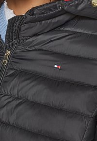 Sort pufferjakke med lynlås og horisontal quilt-design. Har et lille broderet logo i rød, hvid og blå.