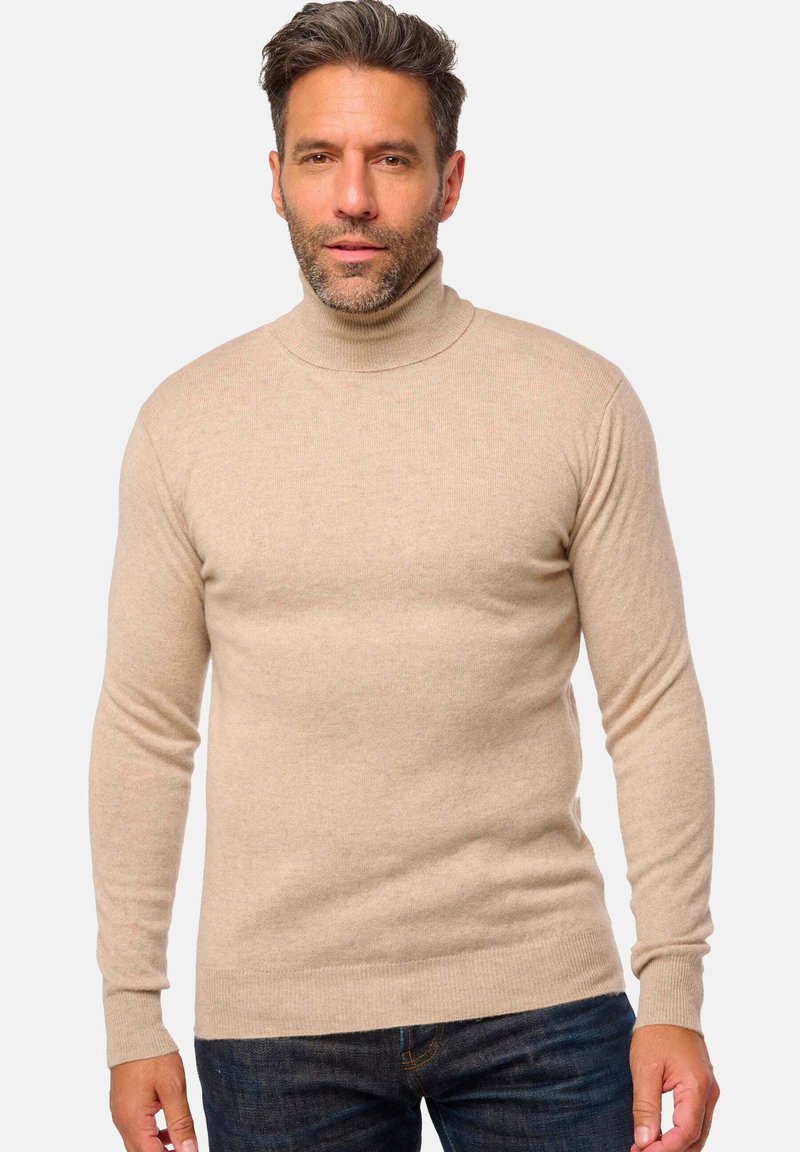 Mann in einem beigen Rollkragenpullover und dunkelblauen Jeans, der vor einem schlichten, hellen Hintergrund steht.
