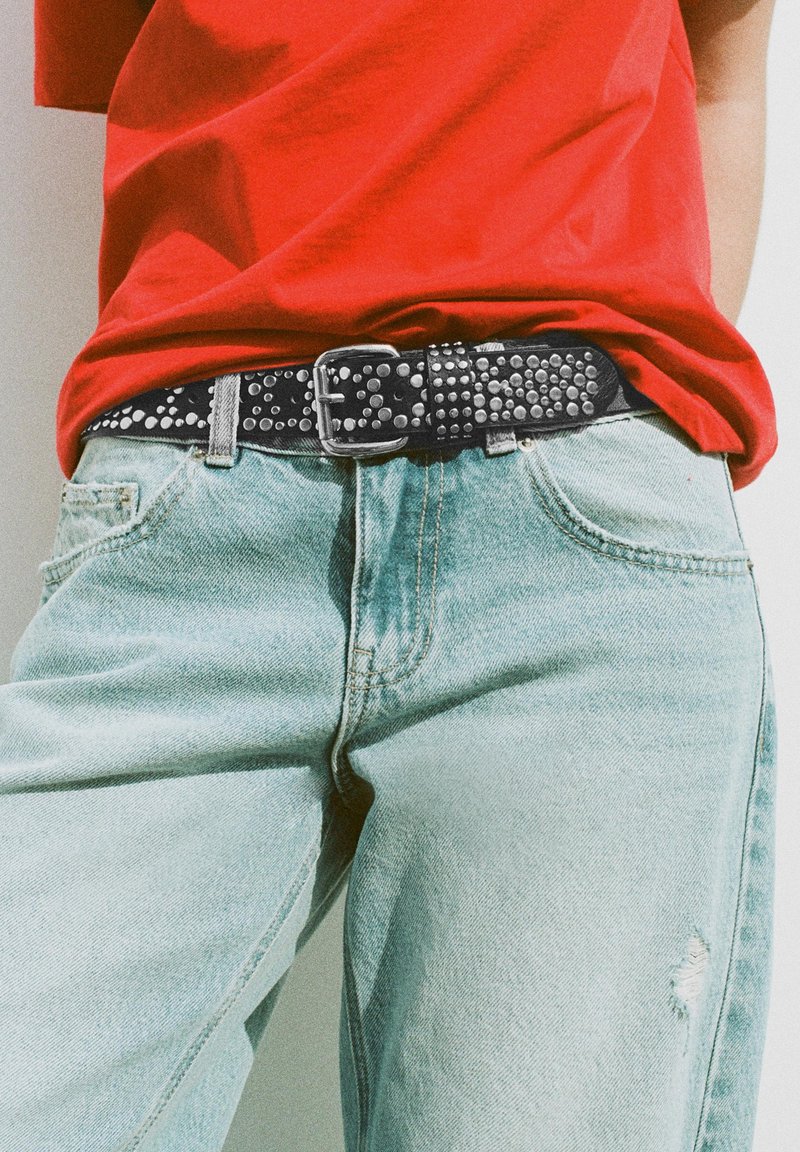 Person trägt hellblaue zerrissene Jeans mit einem schwarzen genieteten Gürtel und einem roten, eingesteckten Hemd.