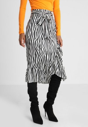 Vrouw draagt een wikkelrok met zebraprint en ruches aan de zoom, gecombineerd met zwarte kniehoge laarzen met hoge hakken en een oranje top met lange mouwen.