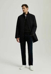 Czarny, wodoodporny płaszcz z wysokim kołnierzem i zapięciem na zamek z przodu, noszony na szaro-swetrowej bluzie i ciemnych dżinsach, połączony z białymi trampkami.