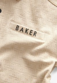 Béžová tkanina s tkaným vzorom, ktorá obsahuje sériu preplietajúcich sa čiar. Na štítku je zobrazené meno "BAKER" čiernym písmom.