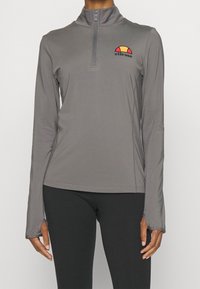 Dlouhý rukáv, šedý sportovní top z hladké látky s půlzipovým límcem. Na přední straně je vyšitý logo v červené, žluté a černé barvě.