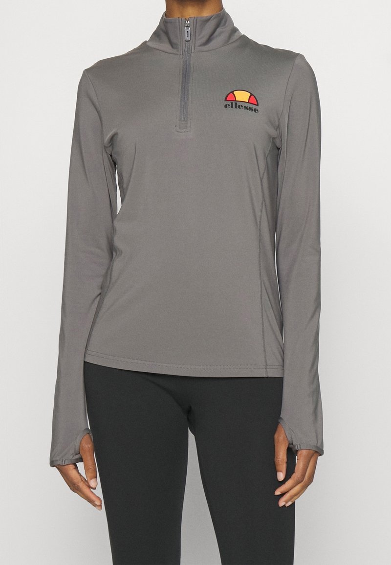 Dlouhý rukáv, šedý sportovní top z hladké látky s půlzipovým límcem. Na přední straně je vyšitý logo v červené, žluté a černé barvě.