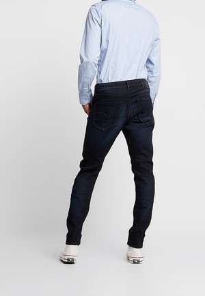 Person trägt dunkelblaue Skinny-Jeans, hellblaues Langarmshirt und weiße Sneaker, steht mit dem Rücken zum Betrachter.