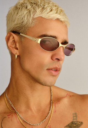 Gros plan sur un jeune homme aux cheveux courts décolorés portant des lunettes de soleil à monture dorée, une boucle d'oreille en or en forme d'anneau, deux colliers en chaîne et un tatouage sur l'épaule.