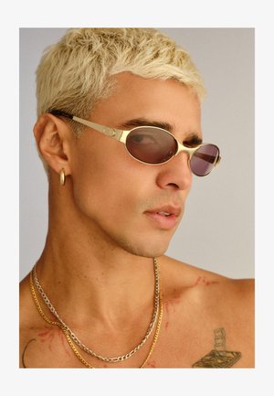 Close-up van een jonge man met gebleekt kort haar die zonnebrillen met een goudkleurig montuur draagt, een gouden oorring, twee kettinkjes en een tatoeage op zijn schouder.