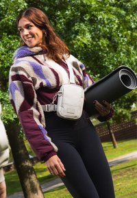 Mosolygó nő a szabadban, színes polárdzsekit, fekete leggingset és keresztpántos táskát visel, kezében egy összegöngyölt fekete jógaszőnyeggel.