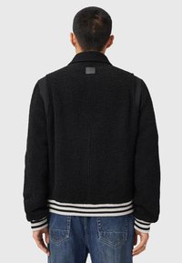 Mand iført sort fuzzy jakke med stribede manchetter og kant, stående med ryggen til, iført blå jeans foran en ensfarvet baggrund.