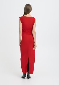 Vestido maxi rojo con detalles de volantes en los hombros, diseño ajustado y una abertura en la parte trasera, combinado con botas negras de tacón. Textura de tela suave.