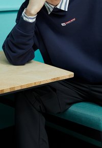 Felpa blu navy con polsini a coste e una patch con logo, indossata sopra una camicia a righe con colletto. Sono visibili pantaloni neri e un tavolo in legno chiaro.