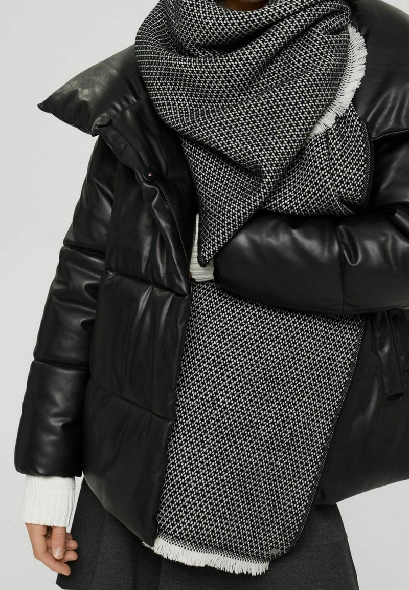 Personne portant une doudoune noire et une grande écharpe à carreaux noirs et blancs avec des bords frangés. Manches de pull blanches visibles.