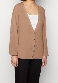 Beige Strickjacke mit V-Ausschnitt, langen Ärmeln und sechs Knöpfen. Sie hat eine gerippte Textur und einen lässigen Schnitt.