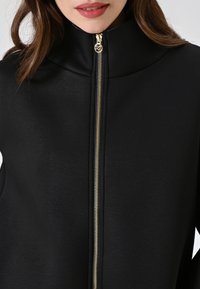Chaqueta negra con cremallera y cuello alto, con una textura suave y detalles en cremallera dorada. El diseño es minimalista y moderno.