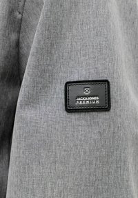 Odev z sivého látkového materiálu s čiernou prišitou štítkom s nápisom "JACK & JONES PREMIUM" a malým geometrickým logom nad textom.