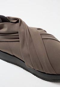 Scarpa in tessuto grigio con design avvolgente, superficie texturizzata e suola in gomma nera con bordo ondulato. Nessun lacci o chiusure visibili.