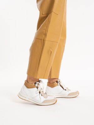 Person wearing cropped tan leather pants and white leather high-top sneakers with beige and gold accents, standing on a white background.