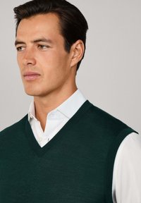 Ciemnozielony dzianinowy sweter w serek noszony na białej koszuli z kołnierzykiem, o gładkiej fakturze i ściągaczach. Zbliżenie koncentruje się na dekolcie.