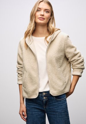 Giacca in pile beige con cappuccio, chiusura con zip, polsini a costine e una consistenza morbida, abbinata a una maglietta chiara e jeans blu scuro.