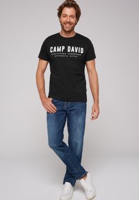 Camiseta de algodón negra con el texto blanco "CAMP DAVID", combinada con jeans de mezclilla azules y zapatillas blancas; atuendo de estilo casual.