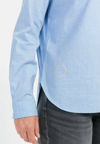 Lichtblauw shirt met lange mouwen, gemaakt van gladde stof. Heeft een gebogen onderkant en een cirkelvormig logo in goud vlakbij de onderkant. Gecombineerd met donkergrijze broek.