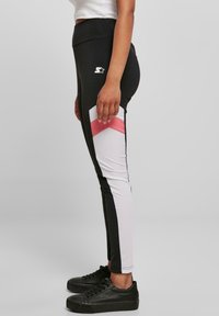 Mustat ja valkoiset leggingsit, joissa on vaaleanpunaisia yksityiskohtia, ja jotka ovat istuvaa muotoilua ja venyviä kangasmateriaaleja. Logo yläreidessä. Yhdistetty mustiin kenkiin.