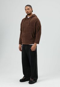 Sweat à capuche en polaire marron avec une poche kangourou, capuche à cordon, associé à un pantalon noir ample et des chaussures noires épaisses.
