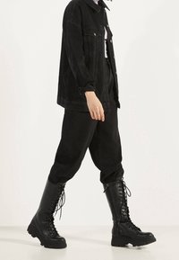 Persona con una chaqueta de mezclilla negra oversize, pantalones negros holgados y botas negras de encaje hasta la rodilla, caminando frente a un fondo blanco liso.
