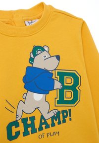 Geel sweatshirt met een afbeelding van een beer in een blauwe hoodie, met de tekst "KAMPIOEN! VAN SPEL" en een grote groene letter "B".