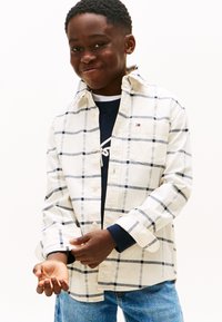 Langærmet ternet skjorte i hvid med marineblå og grå firkanter. Har en knappet krave, venstre brystlomme og opbøjede manchetter over en mørk sweater.