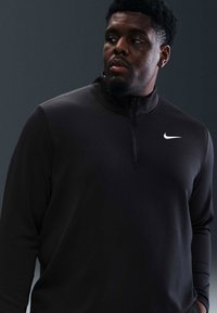 Svart idrettsgenser med kvart glidelås og teksturert stoff, med en liten hvit Nike-logo på venstre bryst. Lange ermer, glatt finish.