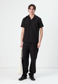 Cămașă neagră cu mâneci scurte și nasturi, împreună cu pantaloni asortați, confecționați dintr-un material texturat. Are dungi verticale și o croială lejeră. Poartă o geantă tote bej.