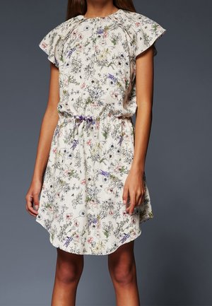 Dziewczyna ubrana w białą sukienkę w kwiaty z falbanami na rękawach i talią ściągniętą fioletową kokardą na ciemnoszarym tle.