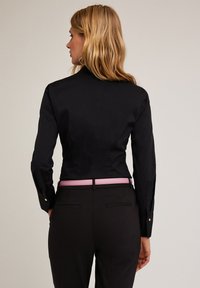 Camisa negra ajustada de manga larga. Presenta un cuello puntiagudo y botones dorados en los puños. Se lleva con pantalones negros a medida y un cinturón rosa.