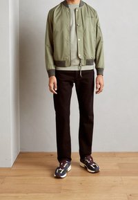 Olivinvihreä bomber-takki, jossa vetoketjusuljenta, resorit hihansuissa ja harmaa huppari alla. Mustat suoralahkeiset housut ja moniväriset tennarit.