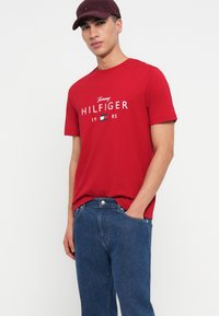 Mladý muž v burgundské čepici, oblečený v červeném tričku s logem Tommy Hilfiger a modrých džínách, s jednou rukou v kapse, stojící na bílém pozadí.
