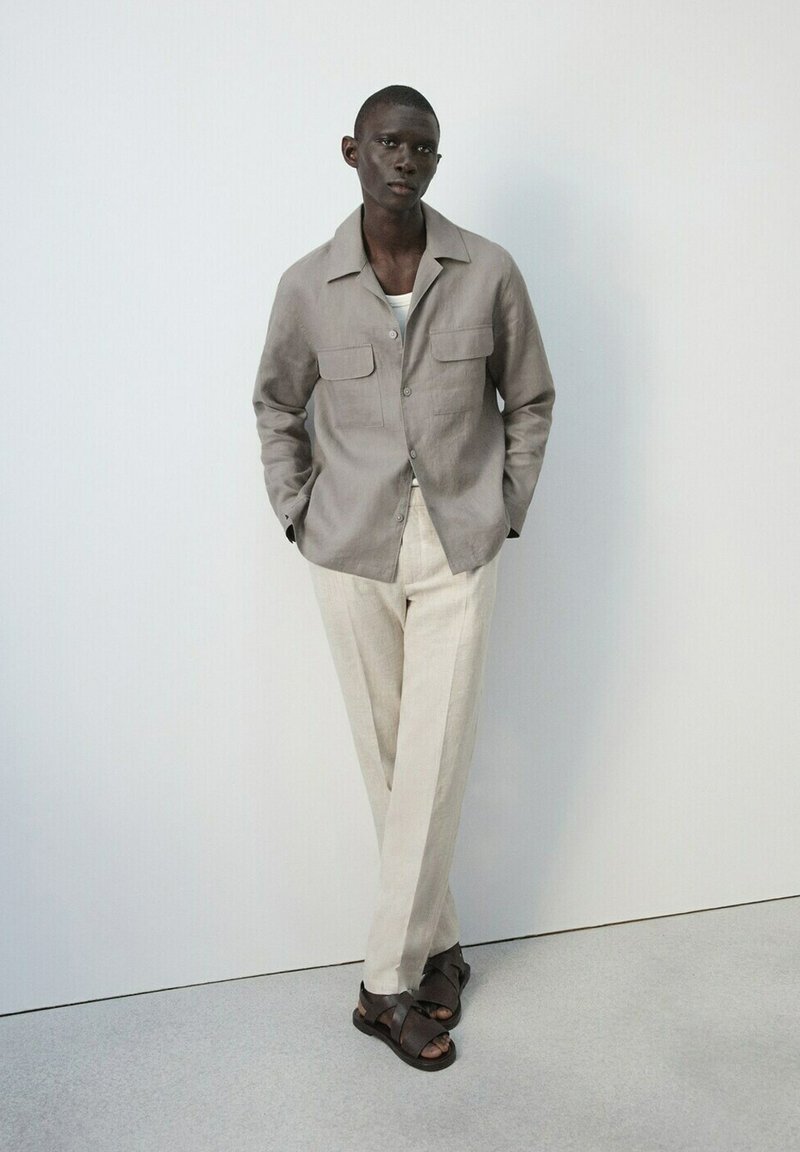 Jeune homme debout, les mains dans les poches, portant une veste beige, un pantalon clair, un sous-vêtement blanc et des sandales marron foncé, sur un fond blanc uni.