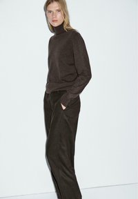 Un pull à col roulé marron en matériau doux, associé à un pantalon sombre. Le pull a des manches longues et un design ajusté, tandis que le pantalon présente des poches latérales.