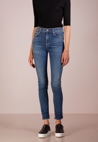 Skinny jeans in medium wash denim, featuring a high waist, distressed accents, and frayed hems, paired with black slip-on sneakers.