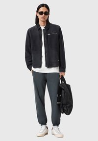 Zwarte suède bomberjack met ritssluiting, witte T-shirt, grijze joggers en witte sneakers. Accessoire: zwarte gestructureerde rugzak.