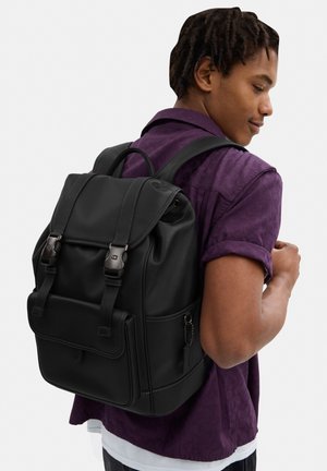 Nuori mies, jolla on päällään tummanvioletti paita, kantaa mustaa nahkaista reppua, joka on kiinnitetty kahdella etuosan soljella.