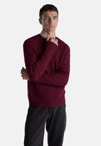 Bordeauxroter Strickpullover mit strukturiertem Rautenmuster, gerippten Bündchen und Saum, kombiniert mit schwarzer Hose. Das Modell steht mit verschränkten Armen.