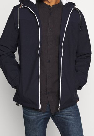 Man wearing a navy zip-up hooded jacket with white lining over a black button-up shirt and blue jeans, hands in jacket pockets.